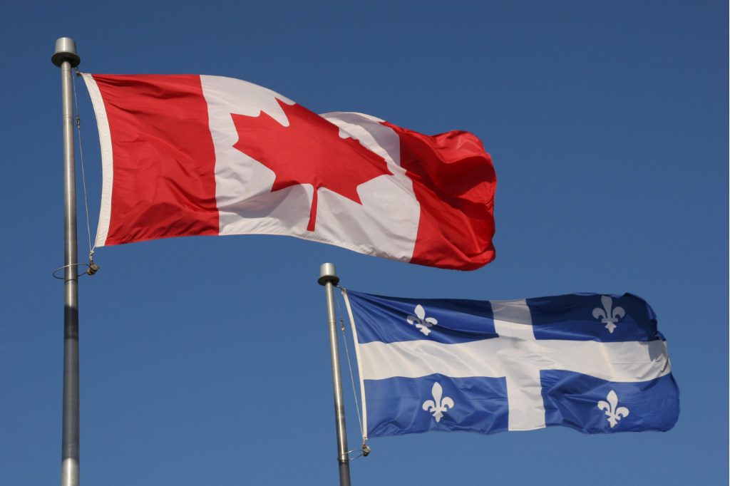 As bandeiras do Canadá e do Quebec estão estendidas em dois mastros diferentes. Atrás dela é possível ver o céu azul.
