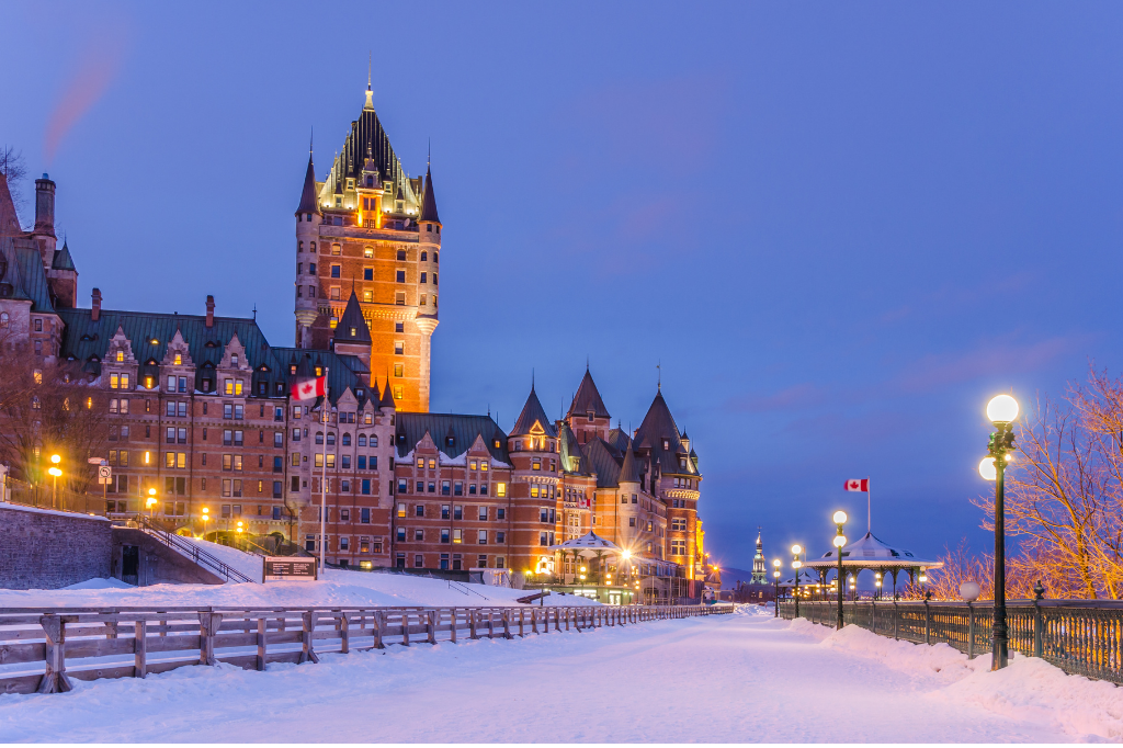 Foto da cidade de Quebeb mostra edifícios iluminados e muito neve no chão. É possível ver a bandeira do Canadá ao fundo.